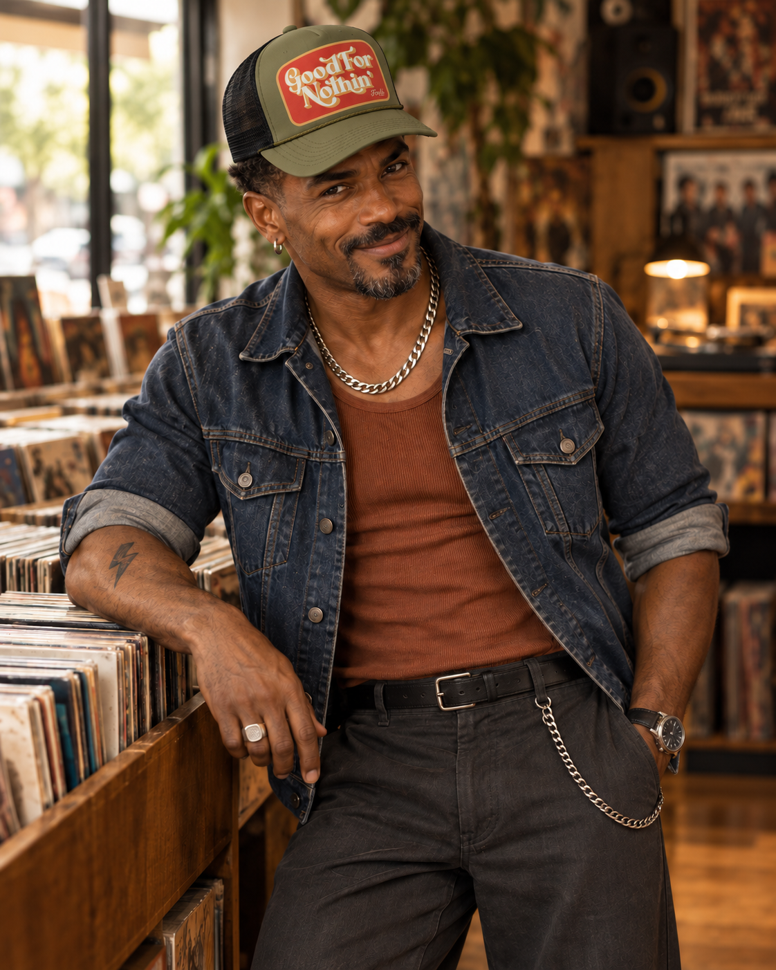 FOOLS Good For Nothin army-green foam and black mesh trucker hat styled with faded denim, rust tank and silver chain in a record shop.