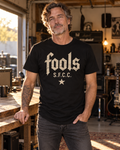 Model concept showing the FOOLS men's black curved-hem tee with distressed bone-colored S.F.C.C. lettering and star artwork.