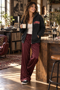 Woman wearing FOOLS Varsity zip hoodie with burgundy satin trousers and silver cross necklace over morning coffee.
