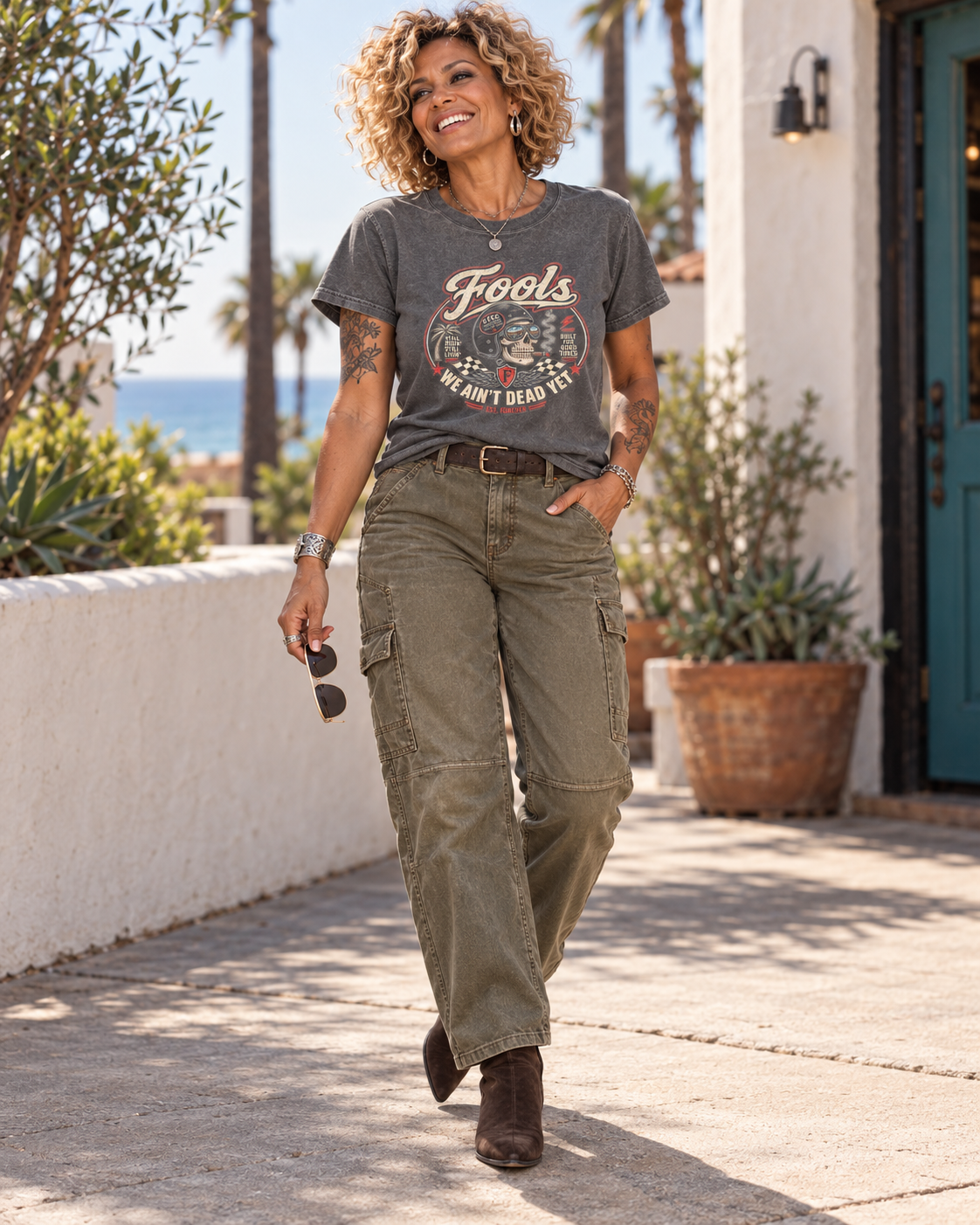 FOOLS Women's Faded We Ain't Dead Yet Tee in faded coal, styled with olive utility pants and brown boots in a coastal courtyard