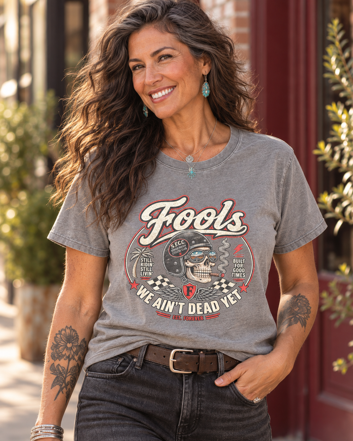 Close-up of the FOOLS Women's Faded We Ain't Dead Yet Tee in faded stone, showing the chest graphic with the dark-haired model, turquoise jewelry and washed black jeans