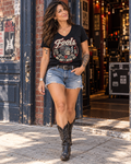 FOOLS Women's We Ain't Dead Yet V-neck Tee in black, styled with vintage denim shorts, a studded belt and black cowboy boots outside a music venue